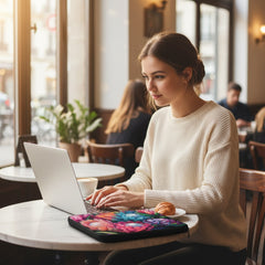 Woman using a laptop in a cafe with a Floral Radiance laptop sleeve by Drese Art on the table